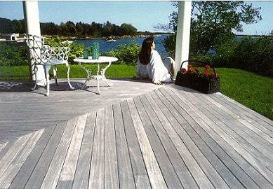 An Ipe deck weathered to a silvery gray patina in a green grass yard on the shorline, with white oron furniture and a woma sitting with her back to the camera on the deck next to a basket of flowers.