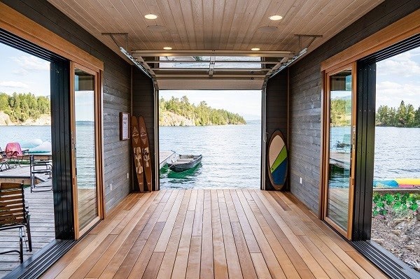 Thermawood Hem-Fir decking in medium brown tones with lighter wood trim and darker stained walls, white overhead door that is open to the water at the end of the deck, with a wood pier on the left of the boathouse outside the doorway, and a sandy area to the right doorway. There is a boat docked on the pier outdside the boathouse overhead door and chairs on the pier. 