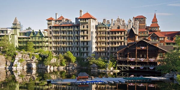 The century old landmark Mohonk Mountain House recently updated the decking on their balconies from concrete to Jatoba wood. In this photo a building that looks like a large castle is reflected in a lake, with rowboats along the pier, green trees, a large rock cliff to the left under one wing of the building., a blue sky with a few clouds, and green, leafy trees.